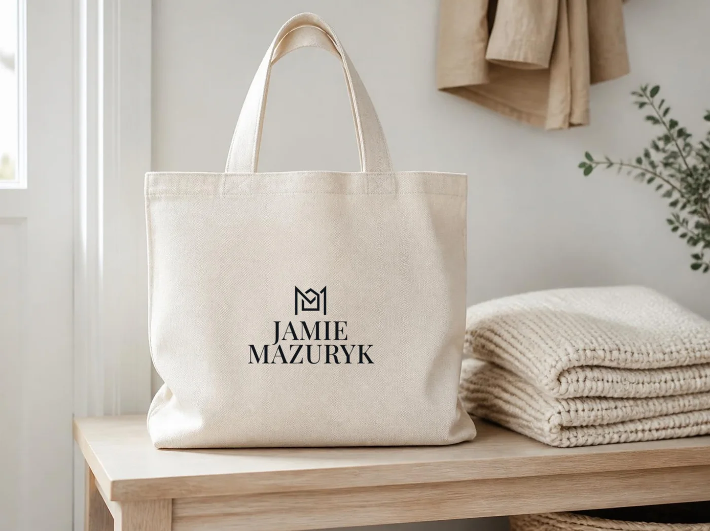 Canvas tote with the Jamie Mazuryk logo next to a folded knit throw